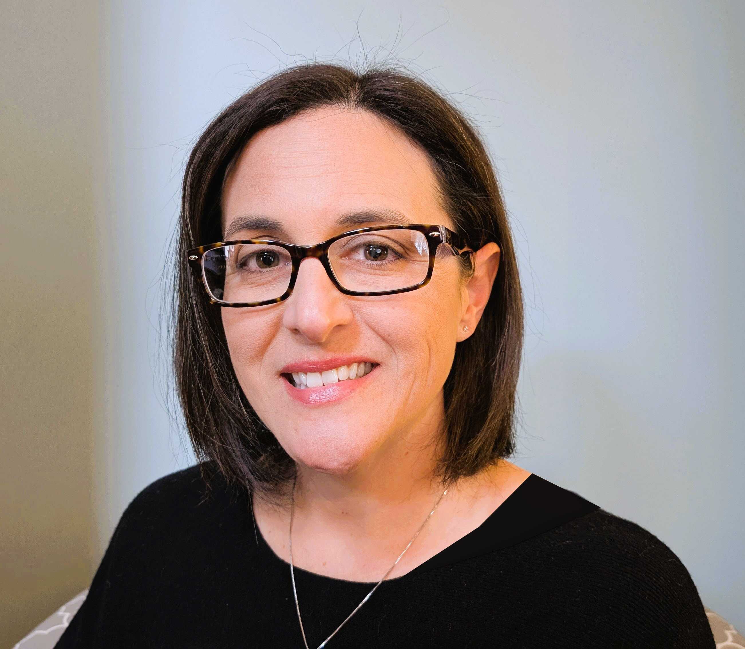 A woman with shoulder-length dark hair and glasses smiles at the camera, wearing a black sweater against a light-colored background.